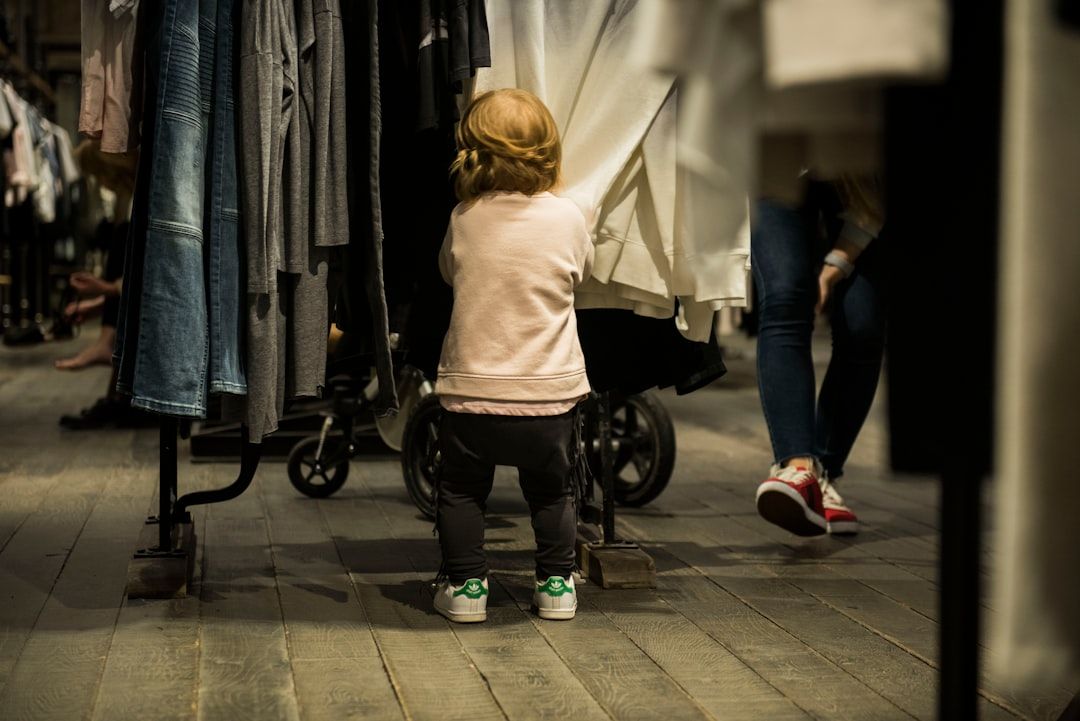 Barn med ryggen mot kameran bland kläder i en butik, med vagn och fötter synliga.